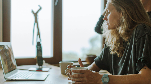 A woman sat at a table with her laptop open, a mug between her palms